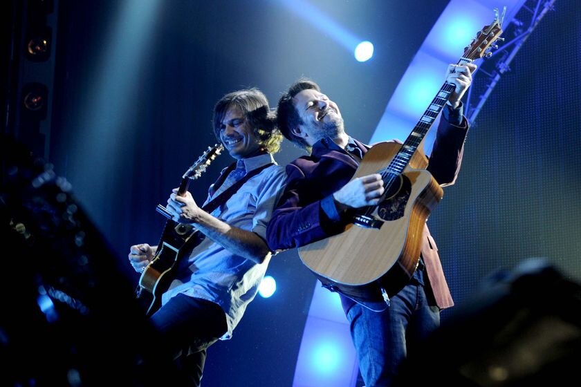 Two guitarists, bathed in blue stage lights, stand back to back, smiling as they play.