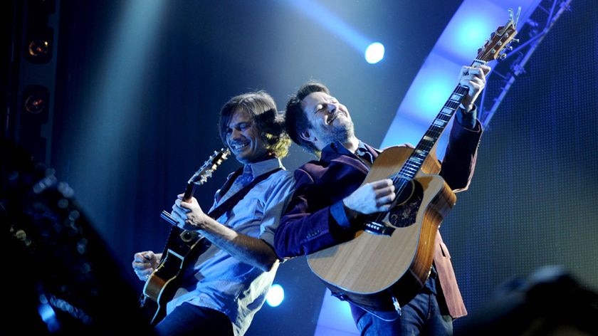 Two guitarists, bathed in blue stage lights, stand back to back, smiling as they play.