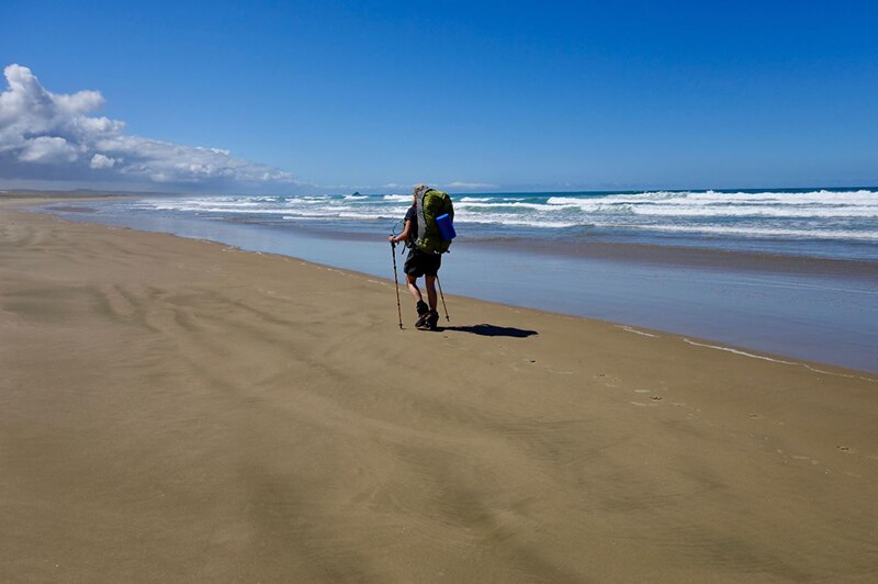 A person with large backpack, shorts, hiking boots and walking sticks walks along a long stretch of empty beach under blue sky.
