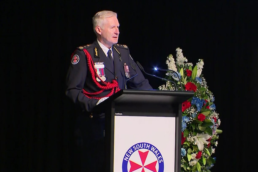 A man in an ambulance officer's uniform speaks at a lectern.
