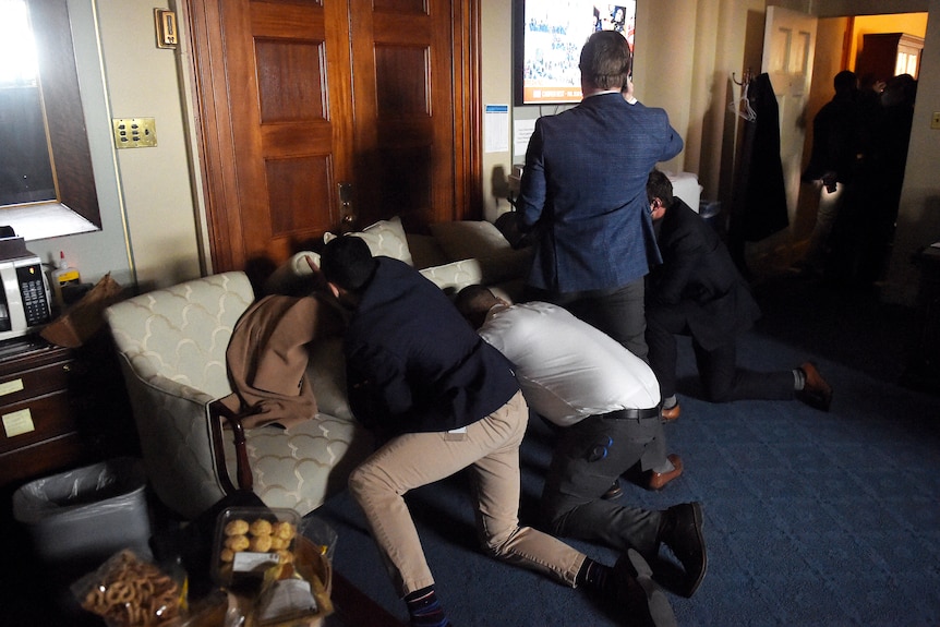 Men crouch behind a couch that has been pushed up against an office door, one stands watching a TV