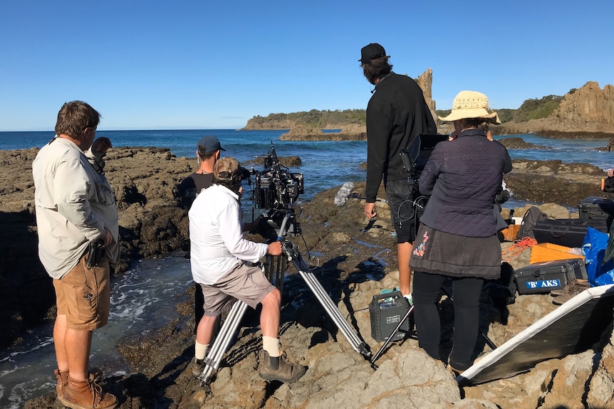 A film crew stand with tech on the rocks looking out to sea. 