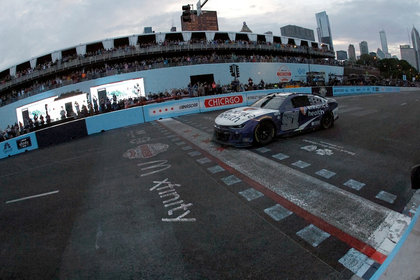 A car crosses the finish line in under grey skies to win a NASCAR race.