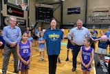A smiling middle-aged woman holds up a blue Townsville T-shirt surrounded by people in gymnasium.