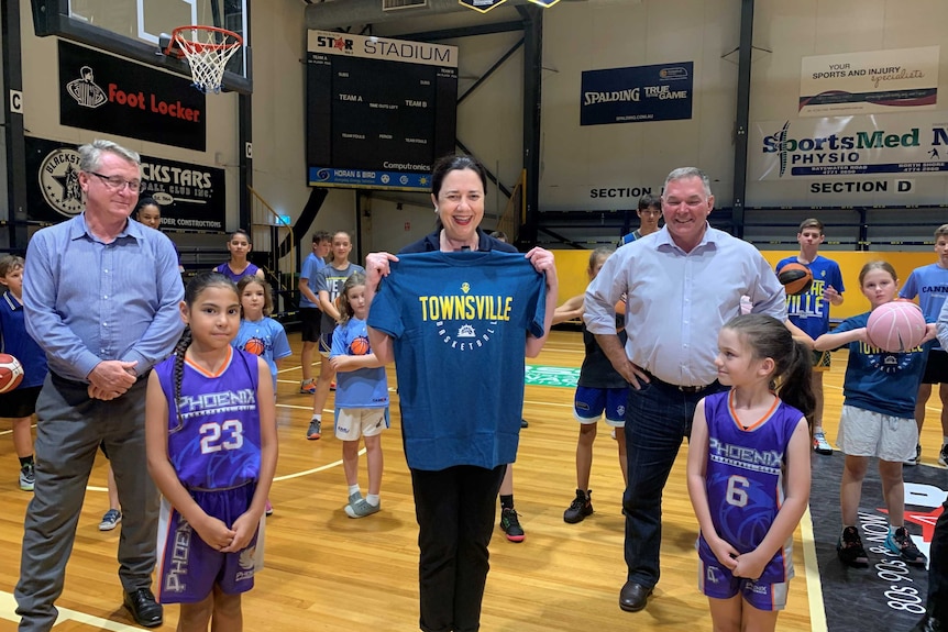 A smiling middle-aged woman holds up a blue Townsville T-shirt surrounded by people in gymnasium.