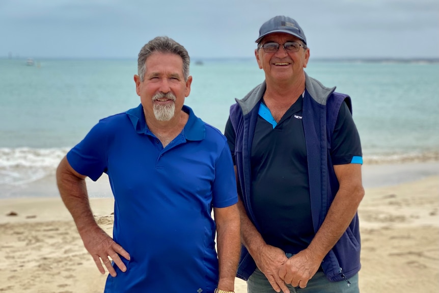 Two men stand facing the camera. The ocean is in the background. 