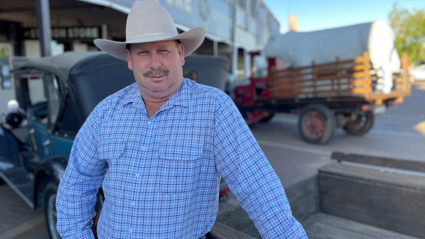 A heavy set man in a blue check shirt and big hat stands in front of two vintage vehicles 