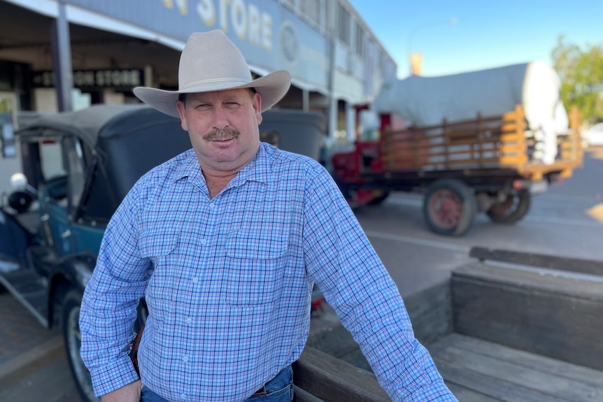 A man in a big hat stands in front of two old cars. 