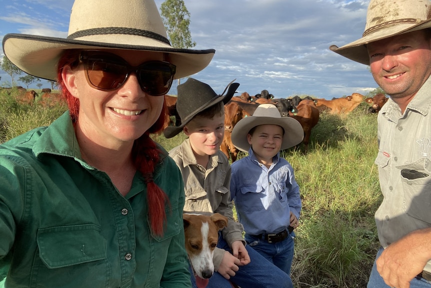 A woman, two small boys and a man wearing hats and work shirts in front of a paddock of cows.