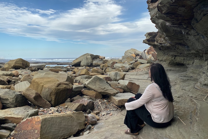 Lisa sits facing away from the camera, sitting on rocks looking towards the ocean