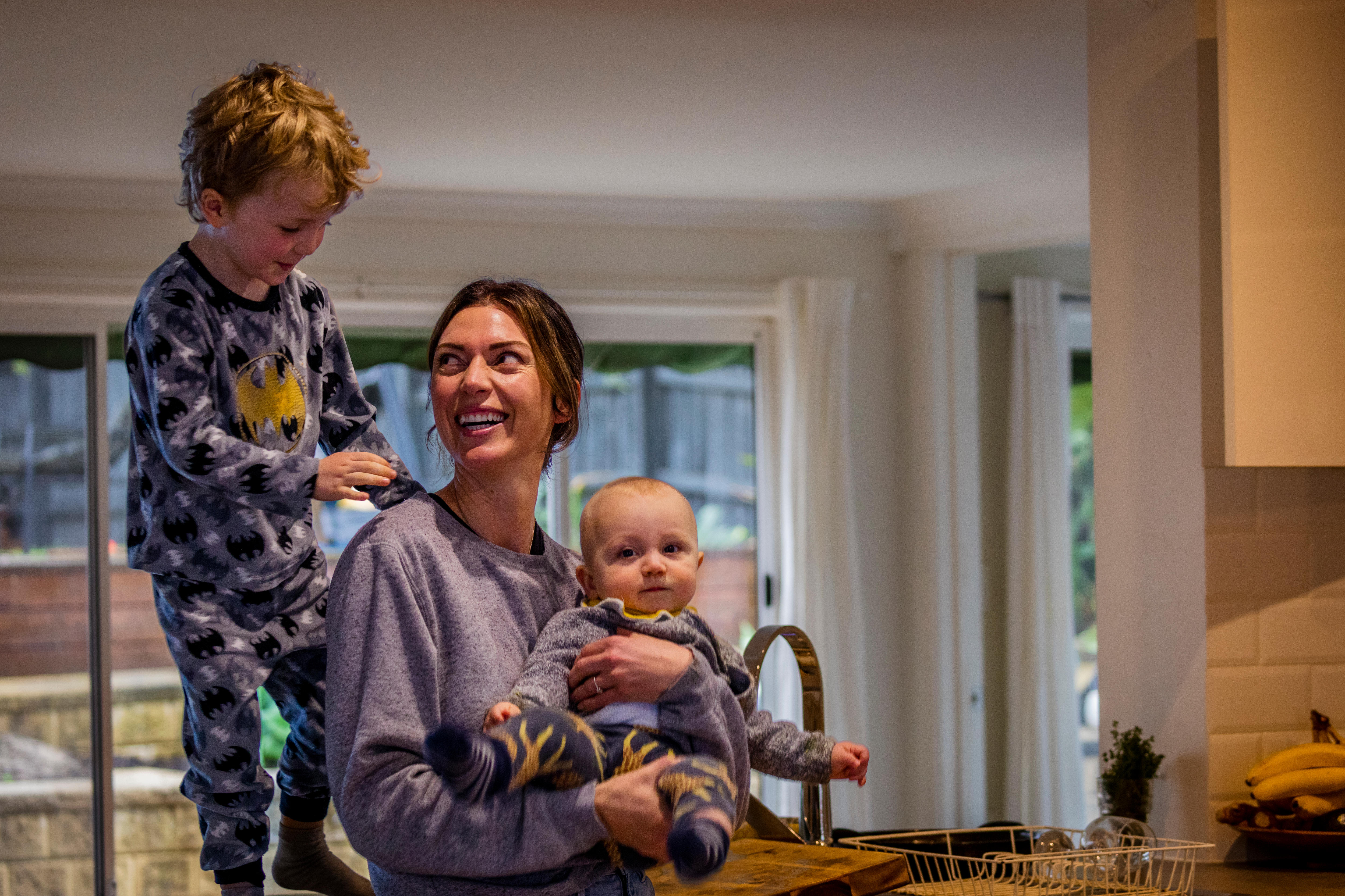Ginelle holds her baby while her young son stands on the kitchen bench behind her smiling at his mum.