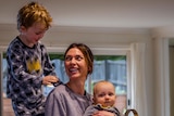 Ginelle holds her baby while her young son stands on the kitchen bench behind her smiling at his mum.