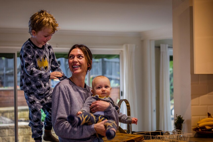 Ginelle sostiene a su bebé mientras su hijo pequeño se para en el banco de la cocina detrás de ella y le sonríe a su madre.