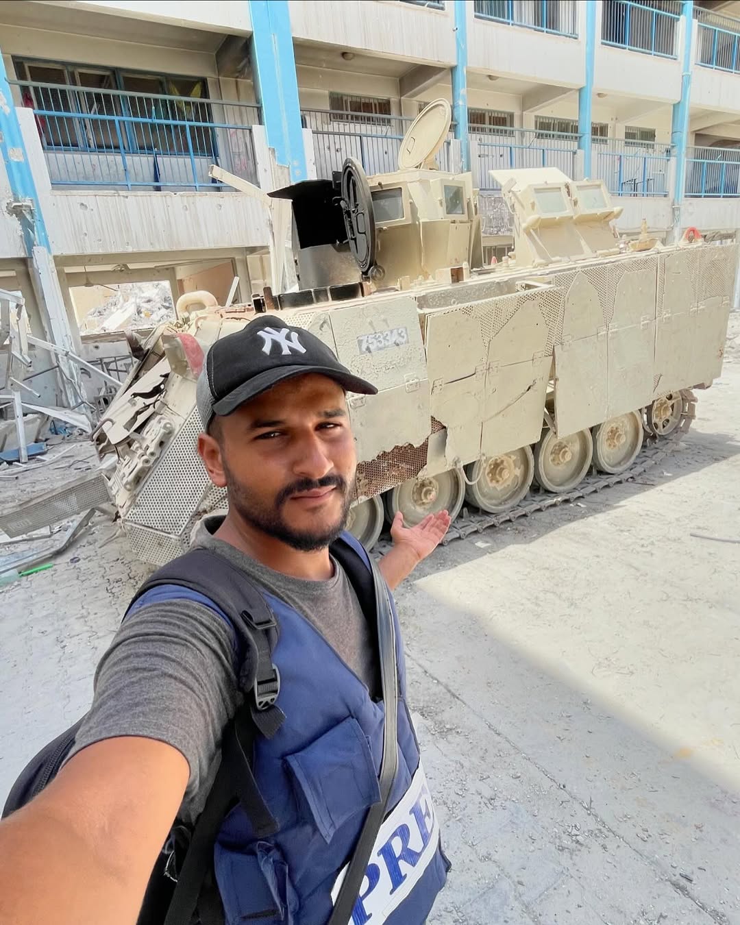A man in a blue vest and black cap stands in front of a tank