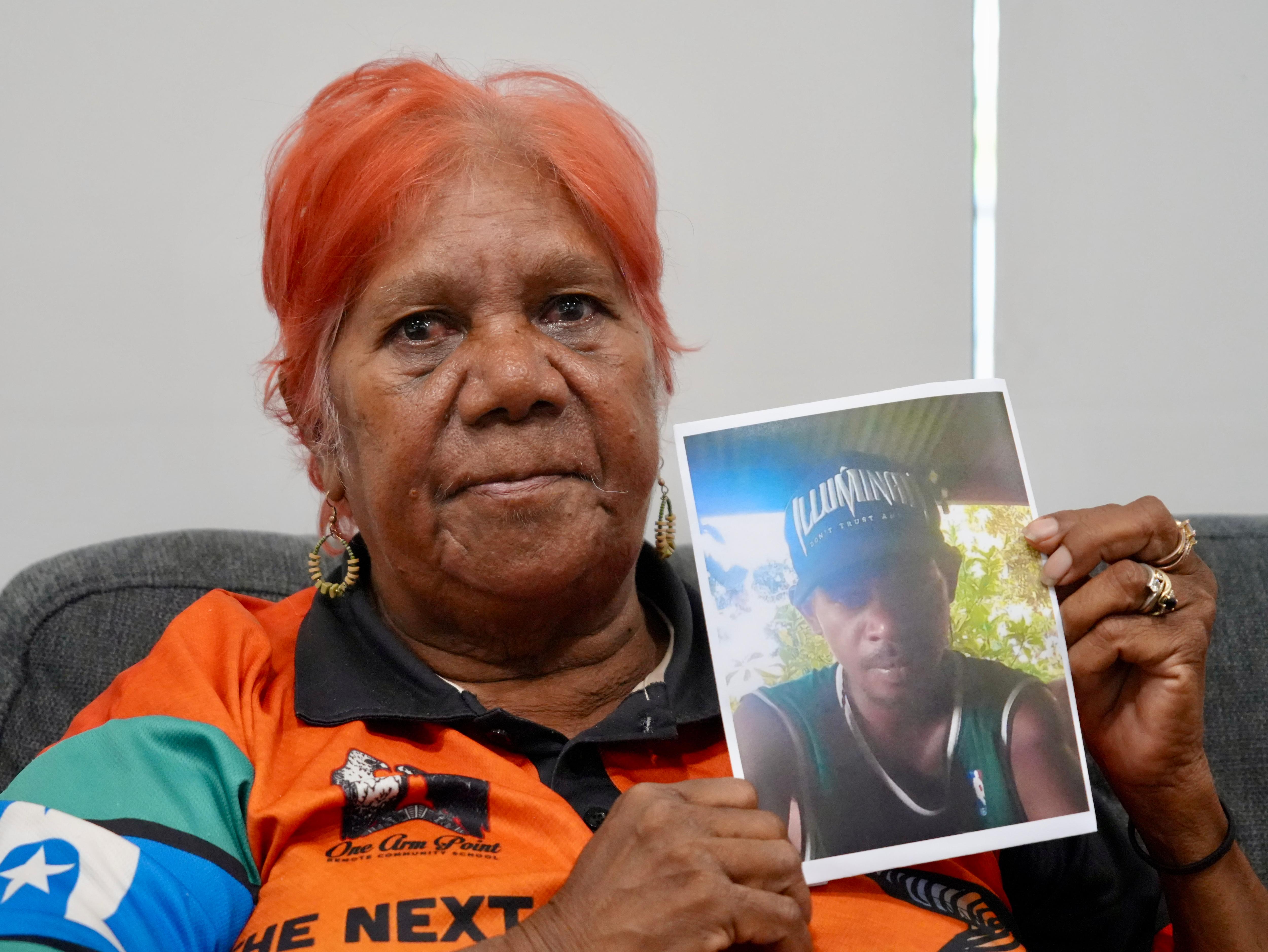 A woman with short hair holds a photo of a young man while looking sad.