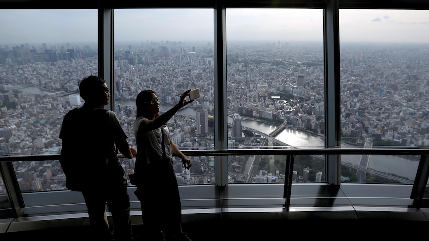 Two people pose for a selfie on an observation deck in a tall building overlooking the Tokyo cityscape.
