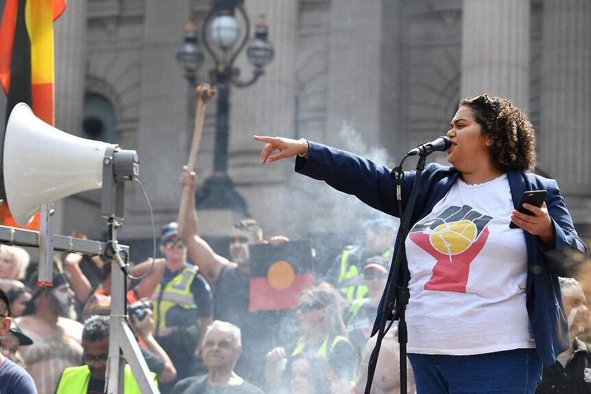 Tarneen Onus Williams is wearing an Aboriginal flag t -shirt and speaks in front of a crowd at people at Melbourne parliament