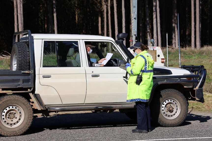 A masked driver of a ute speaks to a police officer wearing a mask and hi vis, writing on a clip board.