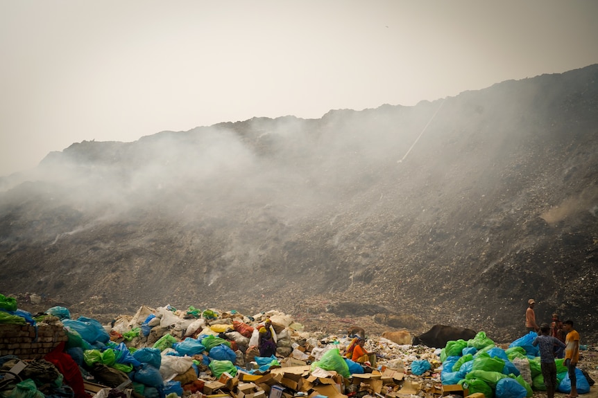 An enormous, towering pile of rubbish surrounded by smoke, while people sit at its base