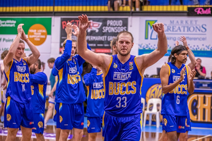 Un homme en uniforme bleu de basket-ball a les bras levés en l'air alors que d'autres applaudissent derrière lui.