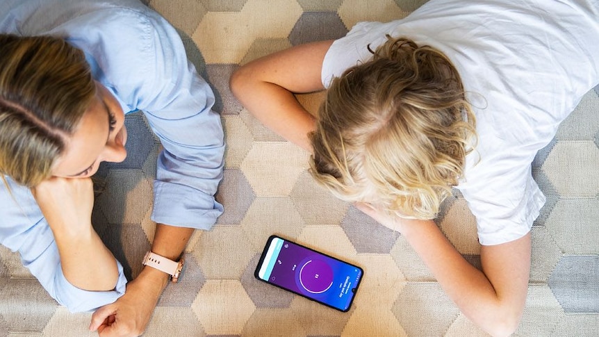 a family of three lies on their tummies on the floor looking at a smart phone that is loading a mindfulness app