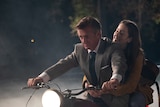 A 60-something man in a suit with slicked back hair and a 20-something woman ride a motorbike, her arms around his waist