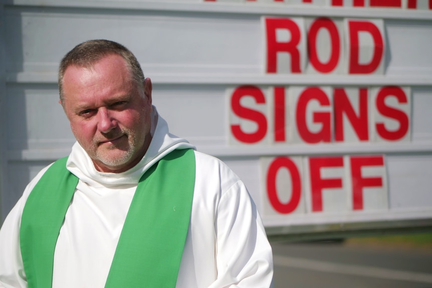 Priest looks at camera with the words Rod Signs off on the sign in the background 