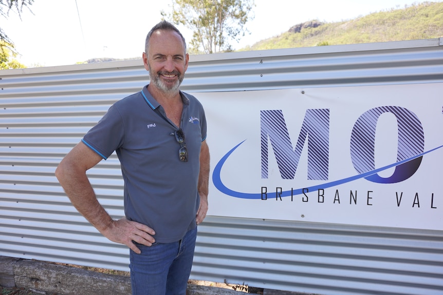 A man wearing a blue polo top stands in front a sign