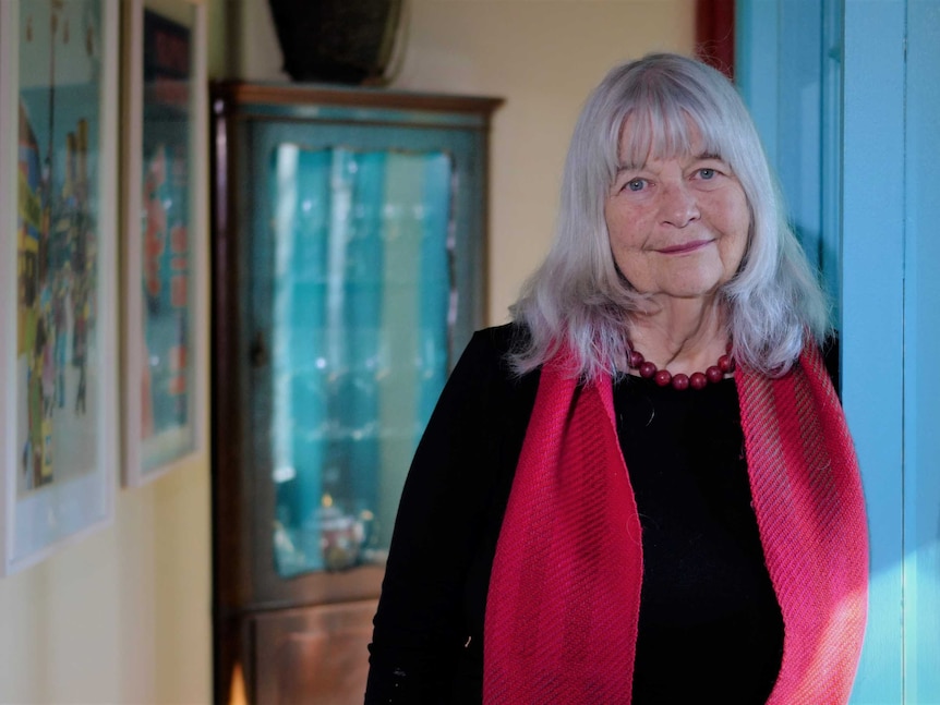 Wendy Bacon stands against a wall looking down the barrel of the camera, with a small smile.