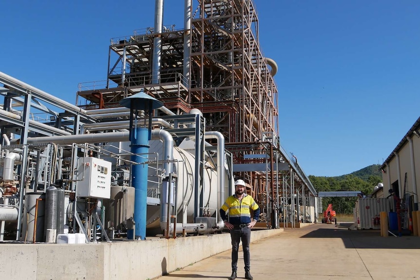 A man in a yellow high-vis shirt and hard hat stands in front a building of metal pipes at biofuels plant.