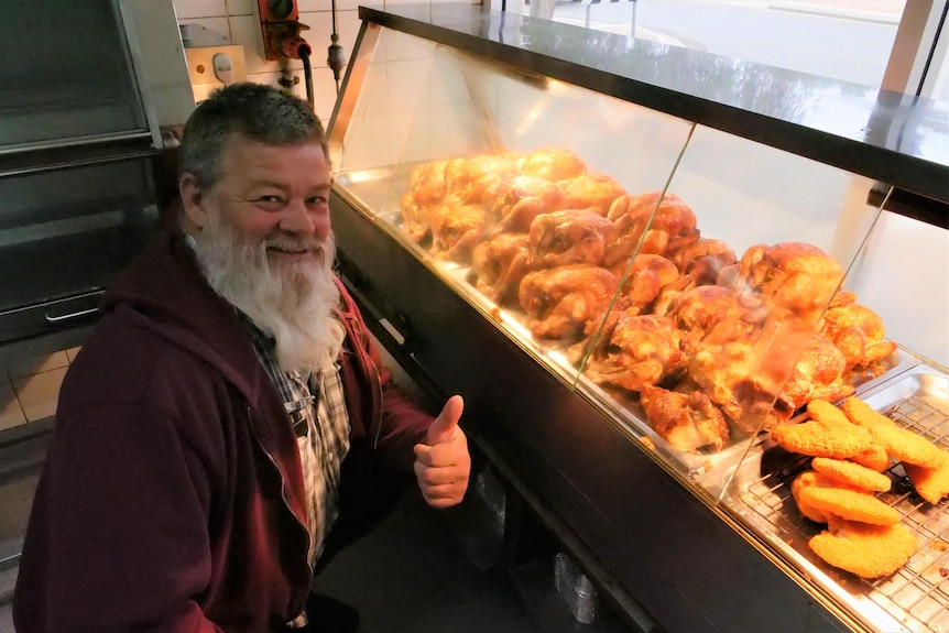 A man with a beard gives a thumbs up in front of a rotisserie containing chickens and potato cakes