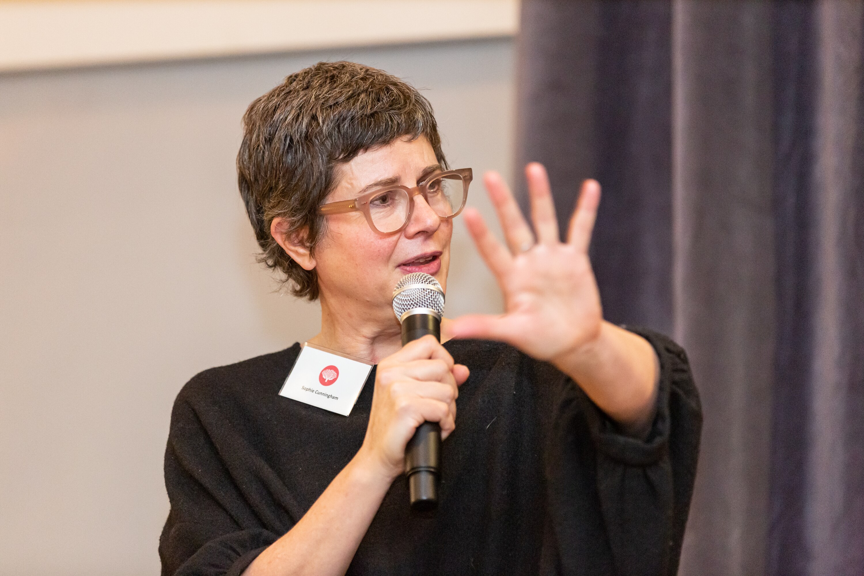 A middle-aged woman with cropped hair, glasses and wearing a black shirt speaking into a microphone, holding her hand out