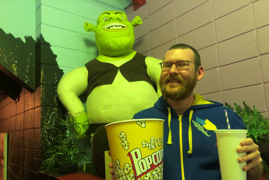 A young man with glasses and a beard stands in front of a Shrek toy with popcorn and a drink.