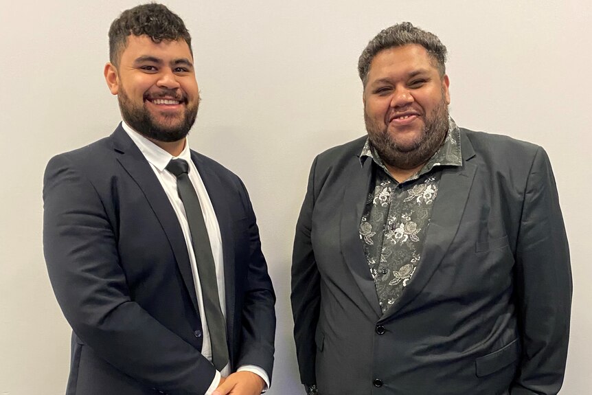Two young Indigenous men standing side by side in suits smiling at the camera 