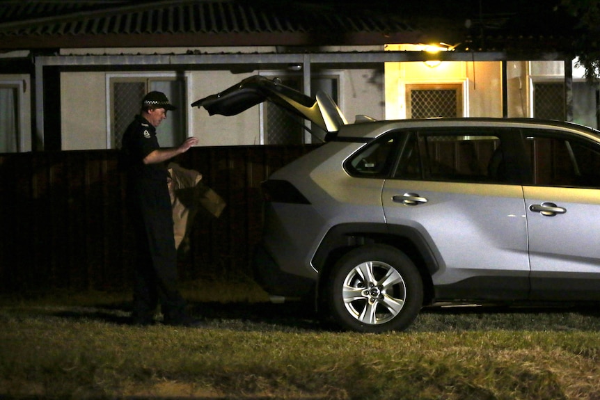 A police forensic officer stands at the back of a silver car holding brown paper bags, outside a house at night.