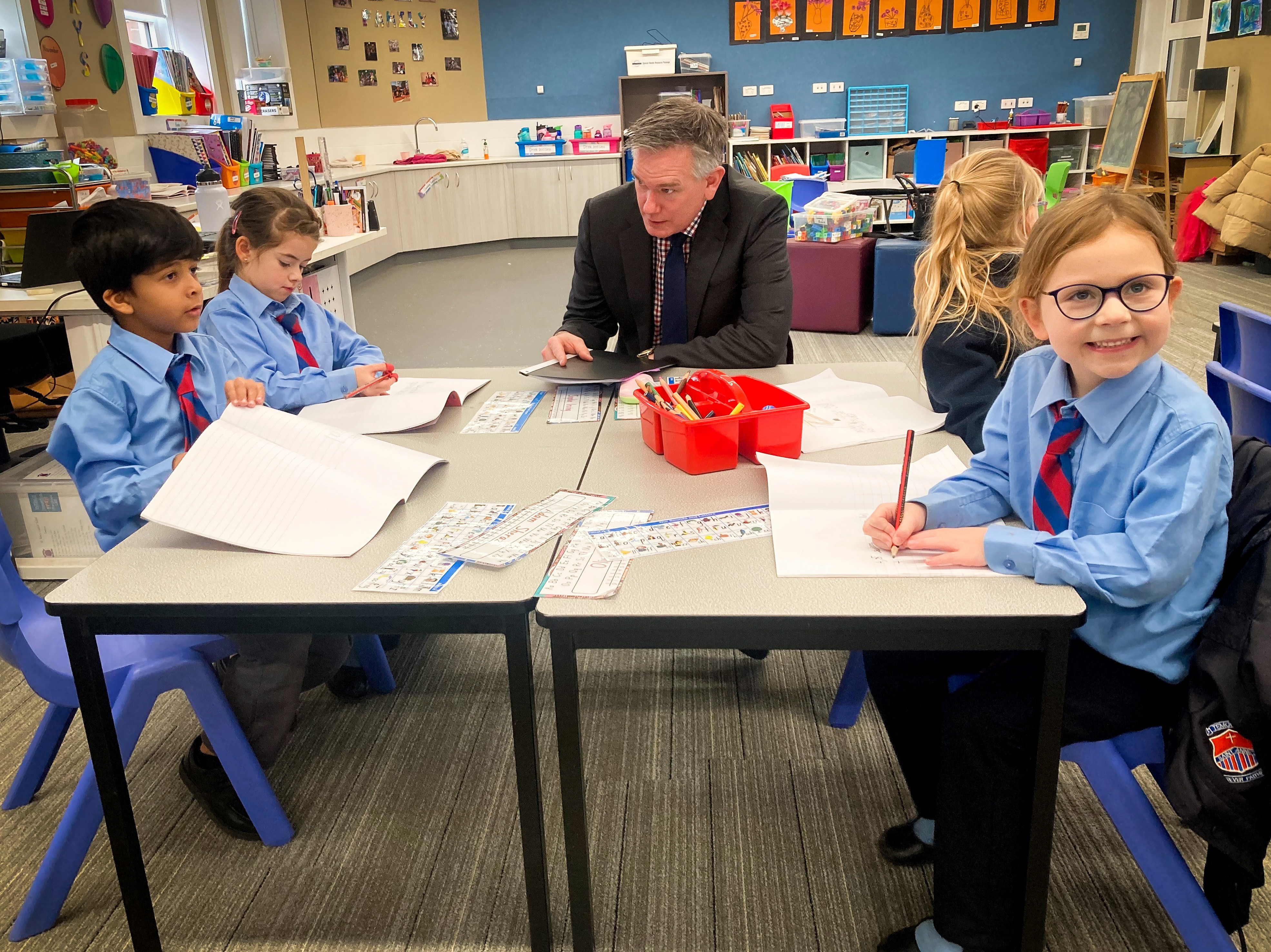 Ross Fox wearing a dark business suit, sitting at a small with four energetic school students writing in their notepads.