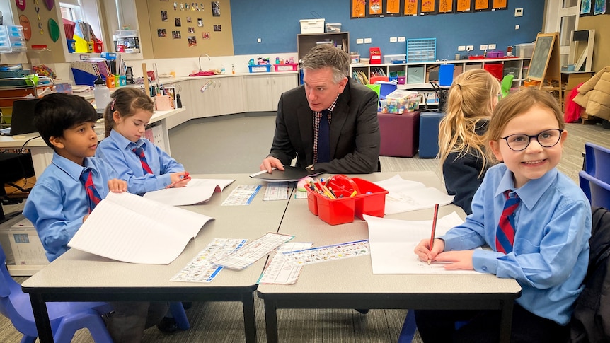 Ross Fox wearing a dark business suit, sitting at a small with four energetic school students writing in their notepads.