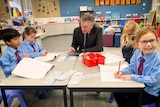 Ross Fox wearing a dark business suit, sitting at a small with four energetic school students writing in their notepads.