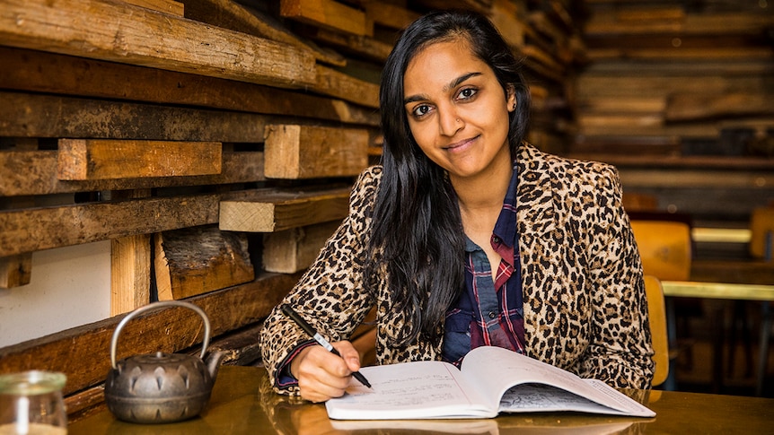 Author Zoya Patel sits at a cafe table writing in a notepad and smiling to camera.
