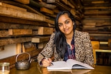 Author Zoya Patel sits at a cafe table writing in a notepad and smiling to camera.