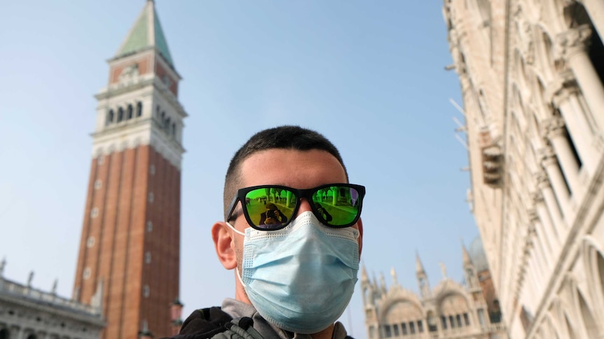 A man with a face mask and sunglasses with Piazza San Marco, Venice, in the background.