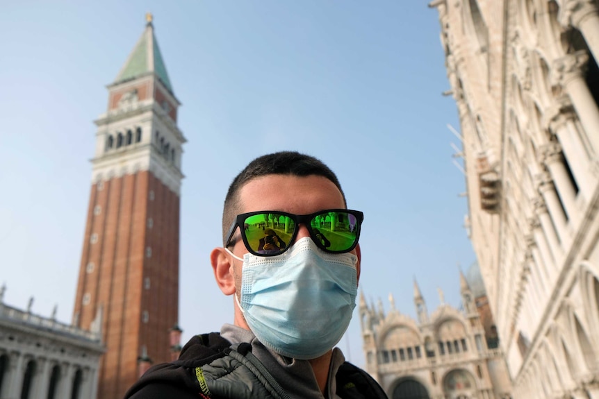 A man with a face mask and sunglasses with Piazza San Marco, Venice, in the background.