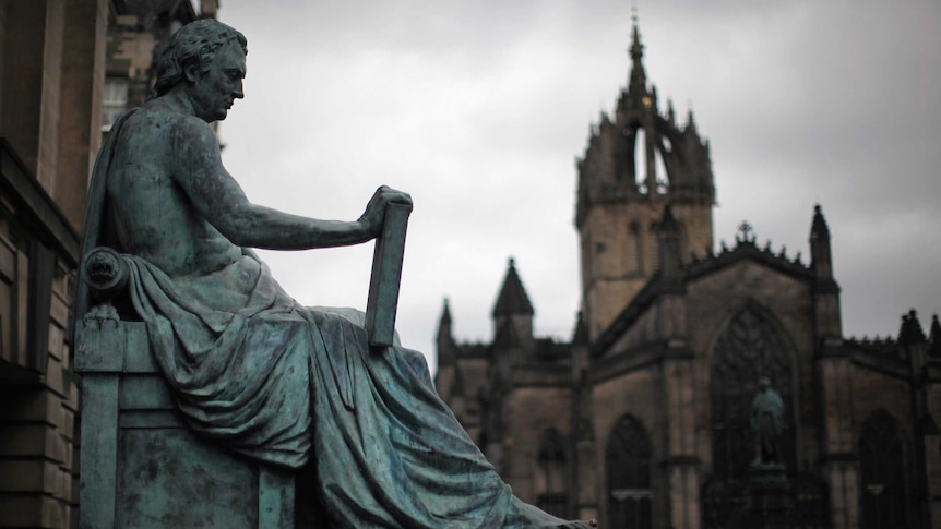 A metal statue of a David Hume holding a book, with cloudy skies and a stone church in the background.