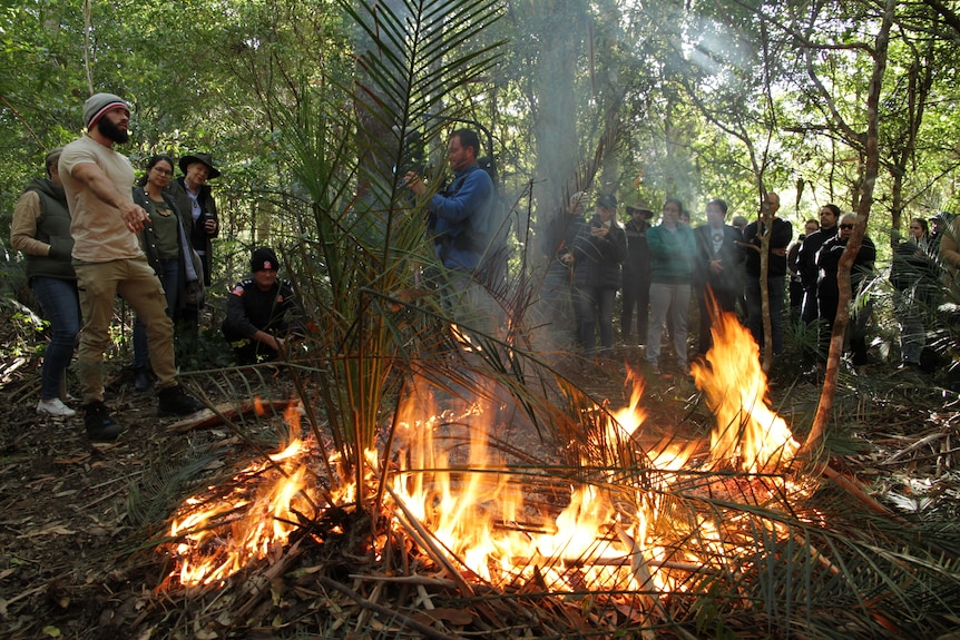 A man points to a shrub on fire, with a crowd of onlookers listening.