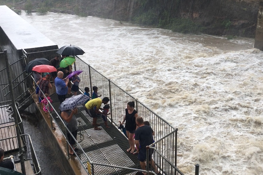 Water rushing down a spillway while onlookers watch on, holding umbrellas.