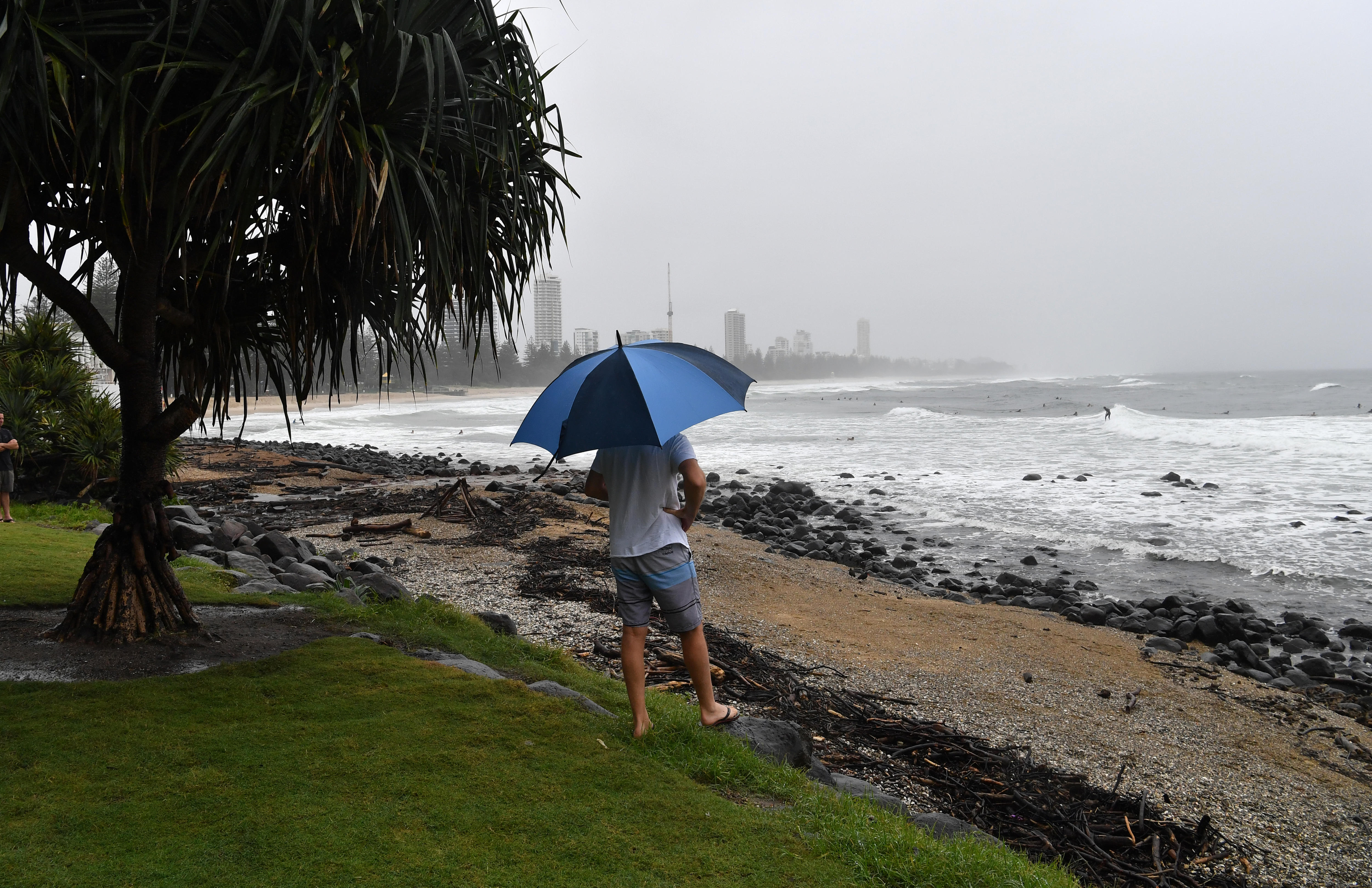 A man holds an umbrella with his back to the camera, overlooking a beach with a city in the background. 