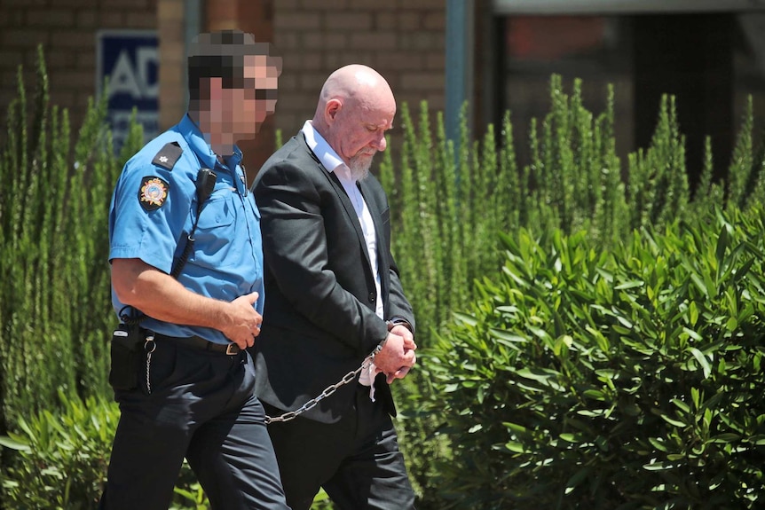 Francis John Wark walks across the road with an officer. They are chained together. The officer's face is blurred.