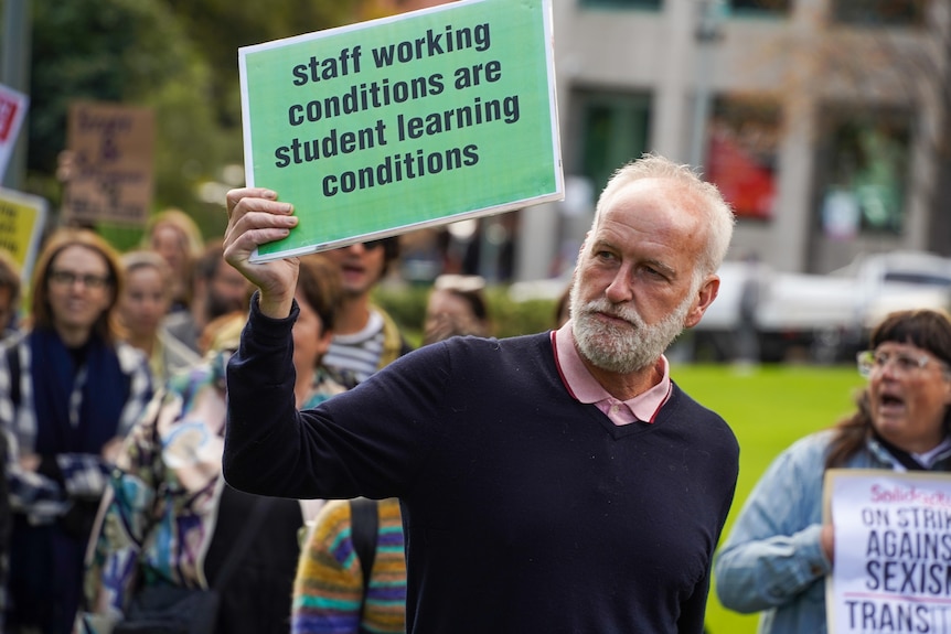 An older man at the university protests holds up a sign which reads "staff working conditions are student learning conditions".