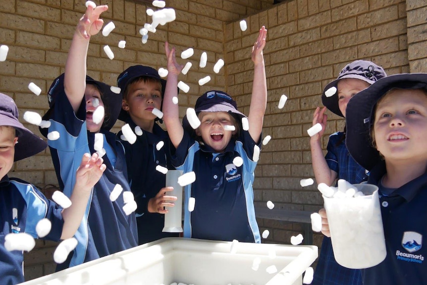 A group of laughing and smiling young students throw styrofoam beads into the air outside their classroom.