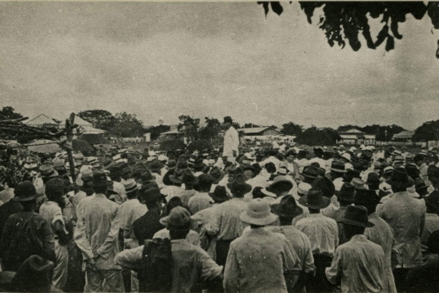 Black and white photo of a group of men standing together with one up higher addressing them.